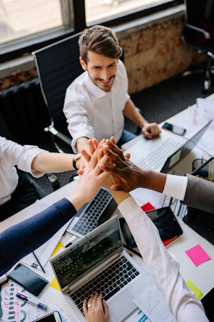 Group of diverse colleagues joining hands around a desk, symbolizing teamwork and unity.