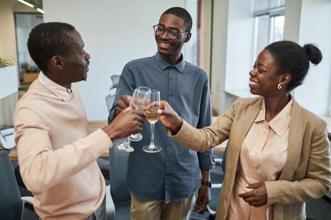 Three colleagues toast with champagne glasses in an office setting, celebrating together.